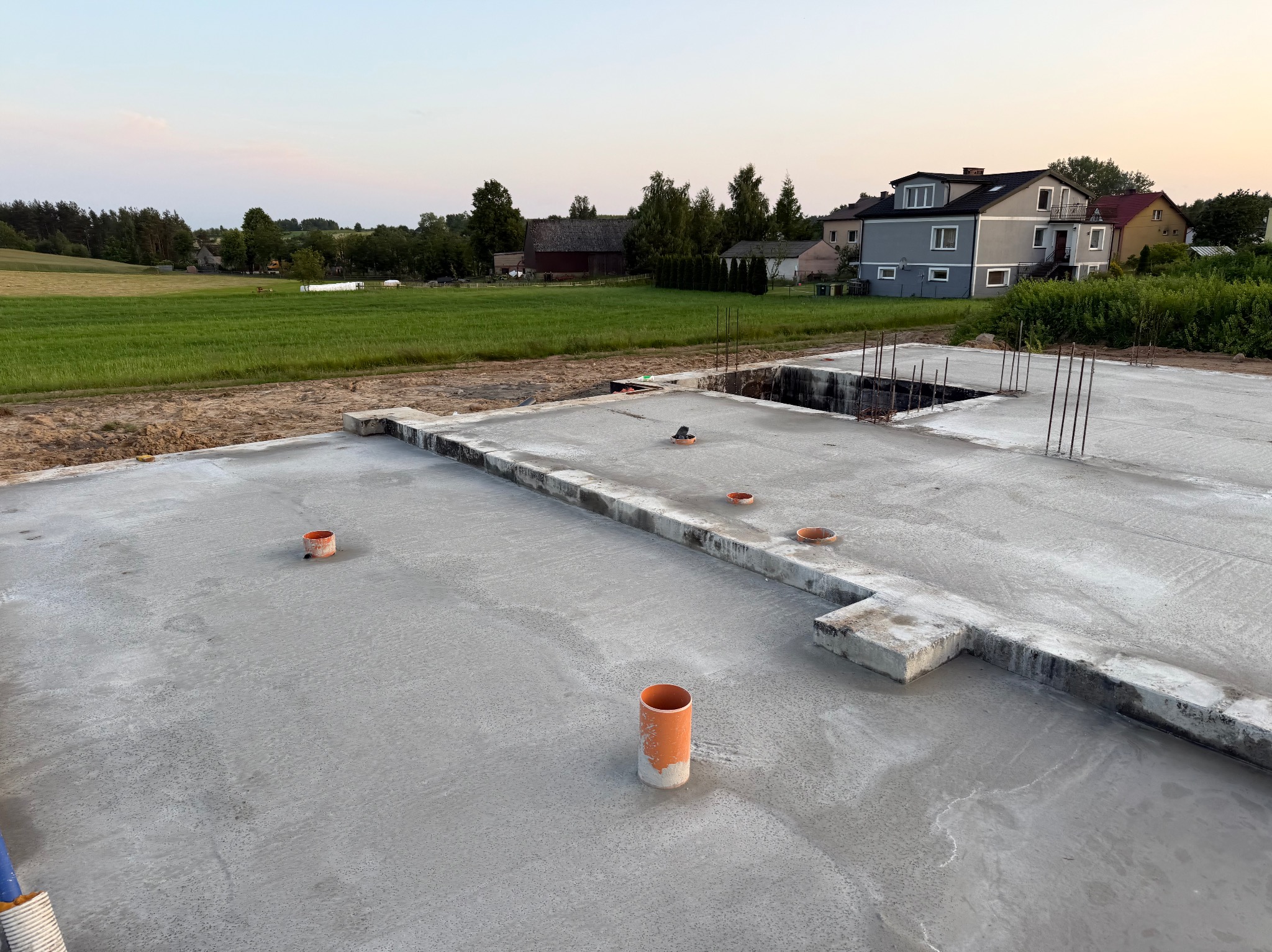 Wylana płyta fundamentowa z wystającymi rurami kanalizacyjnymi i prętami zbrojeniowymi. W tle dom w zabudowie jednorodzinnej i zielone pola.
