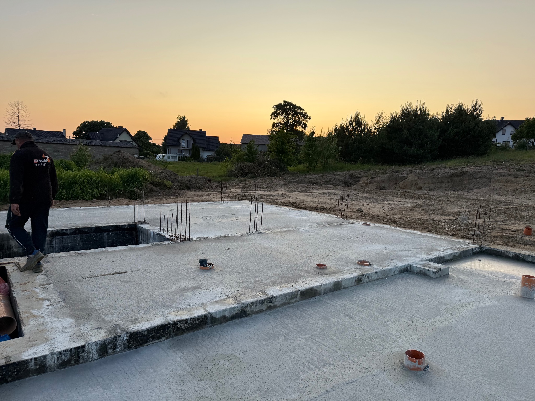 Zalana płyta fundamentowa z wystającymi prętami zbrojeniowymi na tle domów i zachodzącego słońca. Widoczny pracownik w ubraniu roboczym.