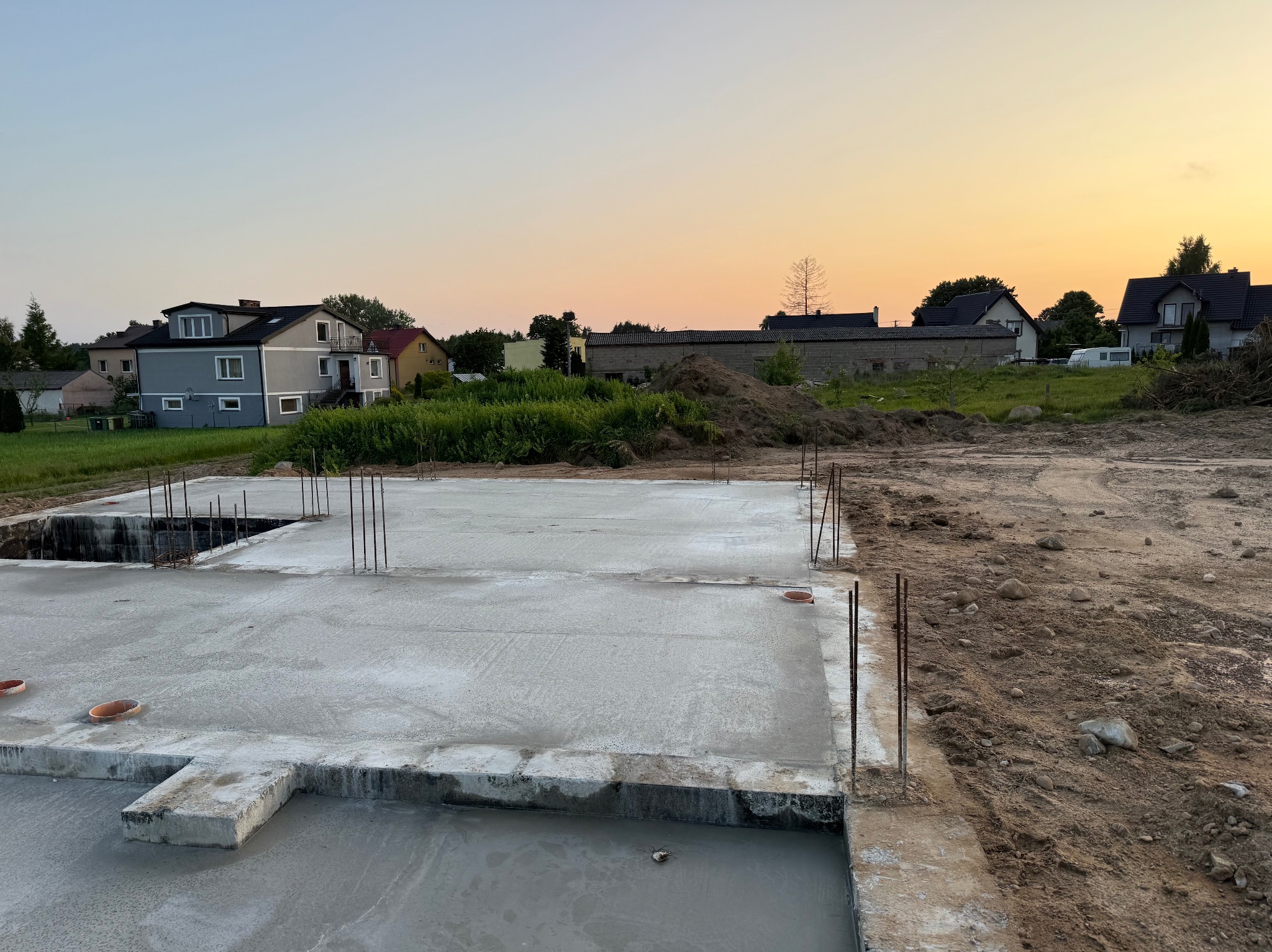 Wylana płyta fundamentowa z wystającymi prętami zbrojeniowymi na tle zabudowy jednorodzinnej i zachodzącego słońca.