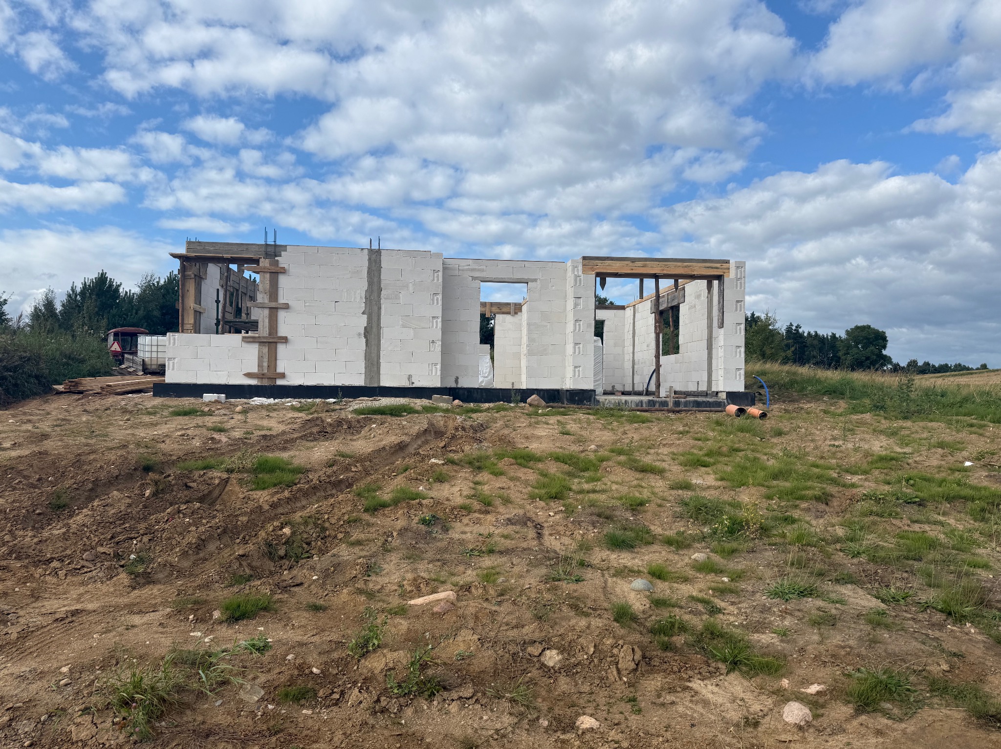 Wznoszenie ścian parterowego domu z białych bloczków, widoczne drewniane nadproża okienne i drzwiowe, w tle błękitne niebo z chmurami i zieleń drzew.