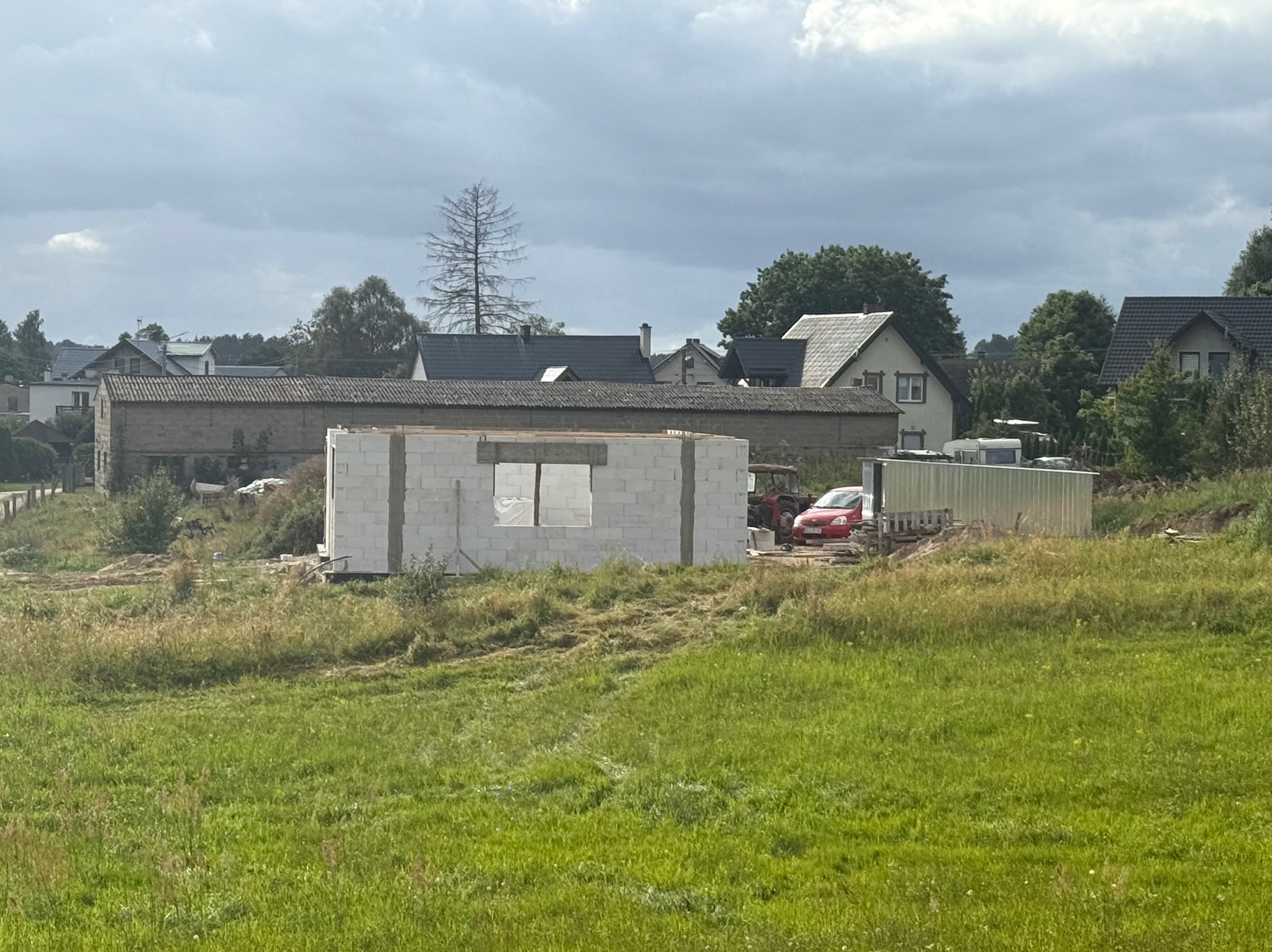 Wznoszenie ścian parterowego domu z betonu komórkowego na tle wiejskiego krajobrazu z zabudowaniami i czerwonym samochodem osobowym.