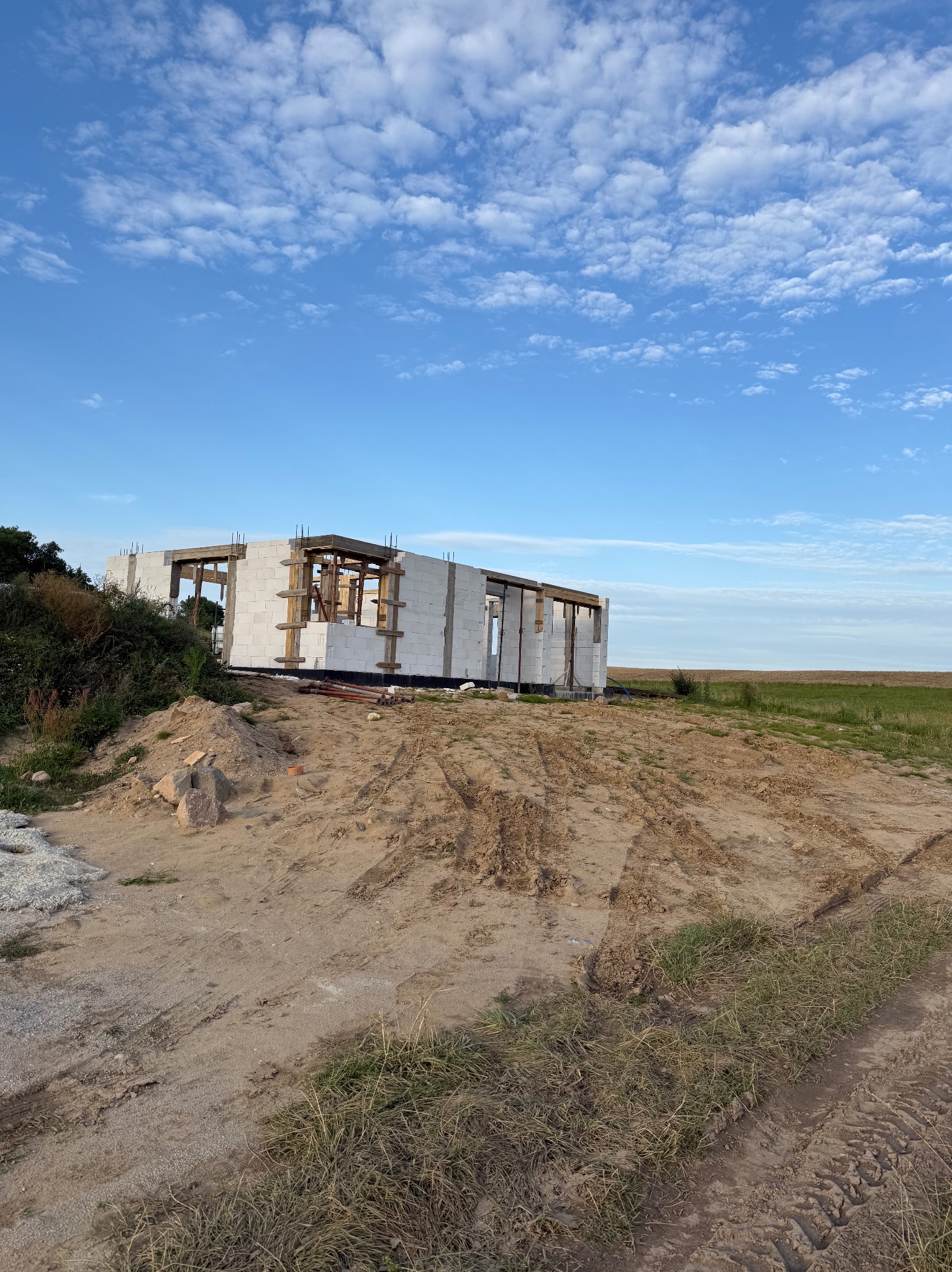 Wznoszony dom z białego bloczka na tle błękitnego nieba z chmurami. Widoczne drewniane elementy konstrukcyjne. Surowy stan, teren budowy z piaskiem i roślinnością.