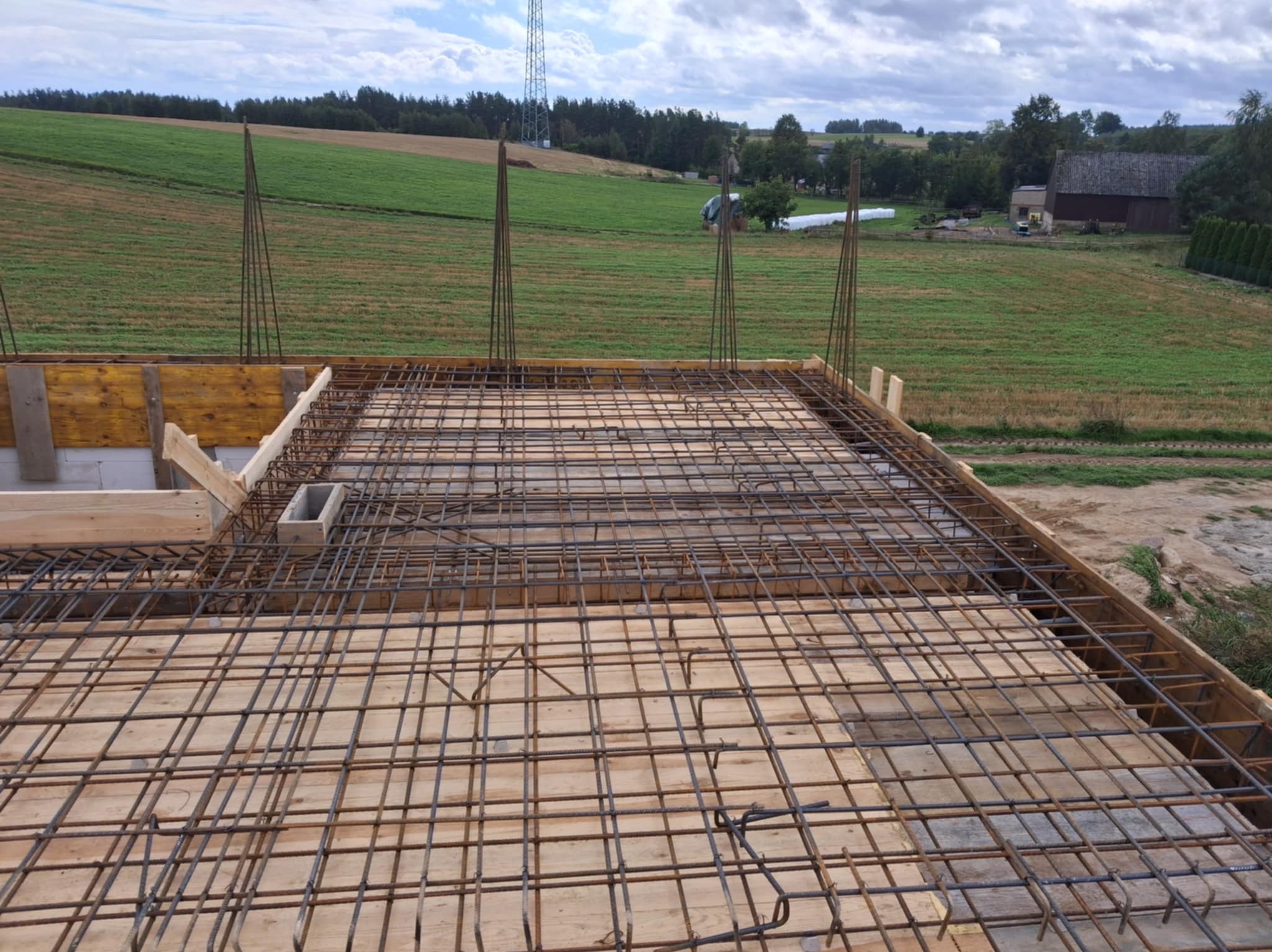 Zbrojenie stropu domu jednorodzinnego z widocznymi prętami i szalunkiem drewnianym na tle zielonych pól i zabudowań gospodarczych w oddali.