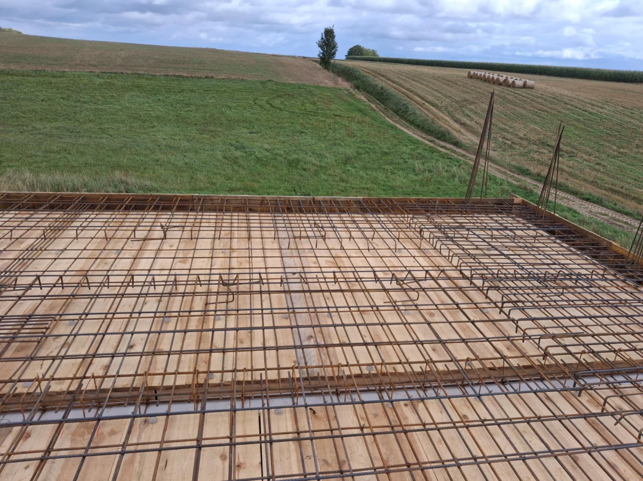 Zbrojenie stropu na deskowaniu z widokiem na pola uprawne i niebo. Stalowe pręty tworzą siatkę, przygotowaną do zalania betonem. Widoczne elementy drewniane szalunku.