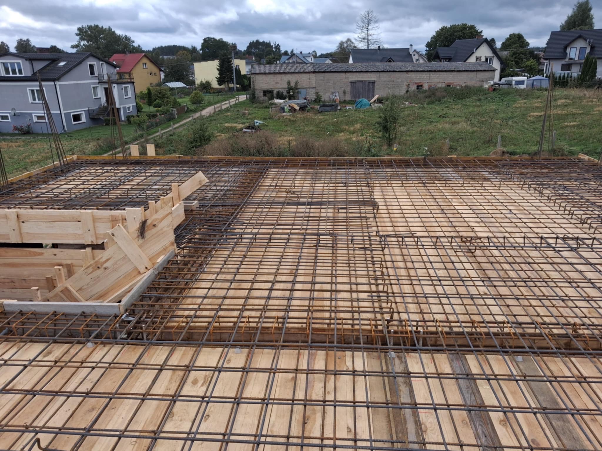Zbrojenie płyty fundamentowej z drewnianym szalunkiem na budowie domu jednorodzinnego. Widok z góry na siatkę zbrojeniową i okoliczne budynki w tle.