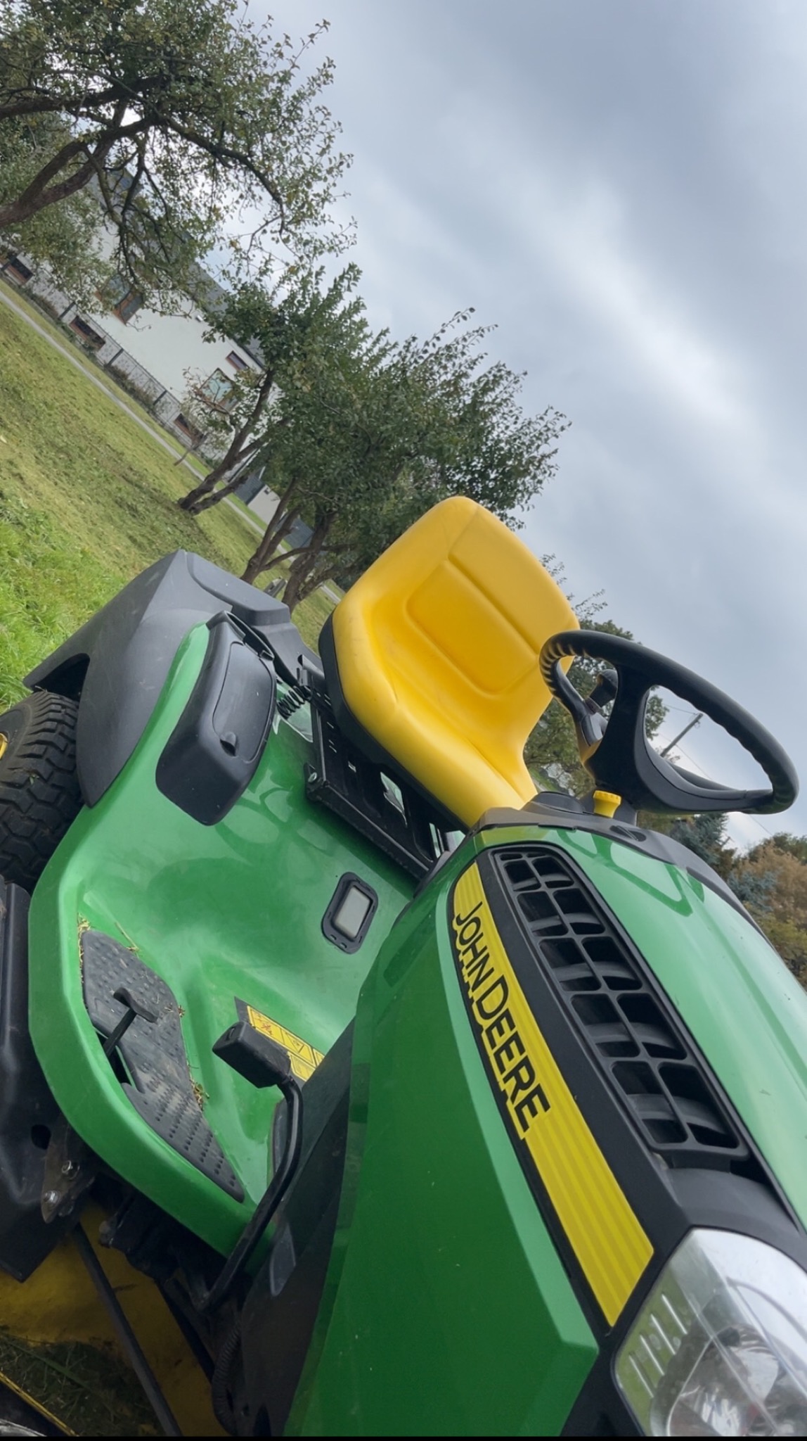 Zielony traktor kosiarka John Deere z żółtym siedzeniem na tle trawnika i drzew w pochmurny dzień. Widok z bliska, lekko obrócony.