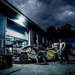 CK Customs - Czarny, oklejony samochód w warsztacie, z otwartą bramą, gdzie widać inne pojazdy. Grupa osób przy aucie, ciemne niebo w tle.