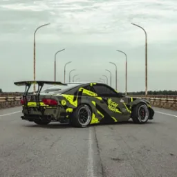 Tuningowany samochód z dużym spoilerem, w barwach khaki i neonowej zieleni, stoi na moście. Widok z tyłu, oświetlenie pochmurne.