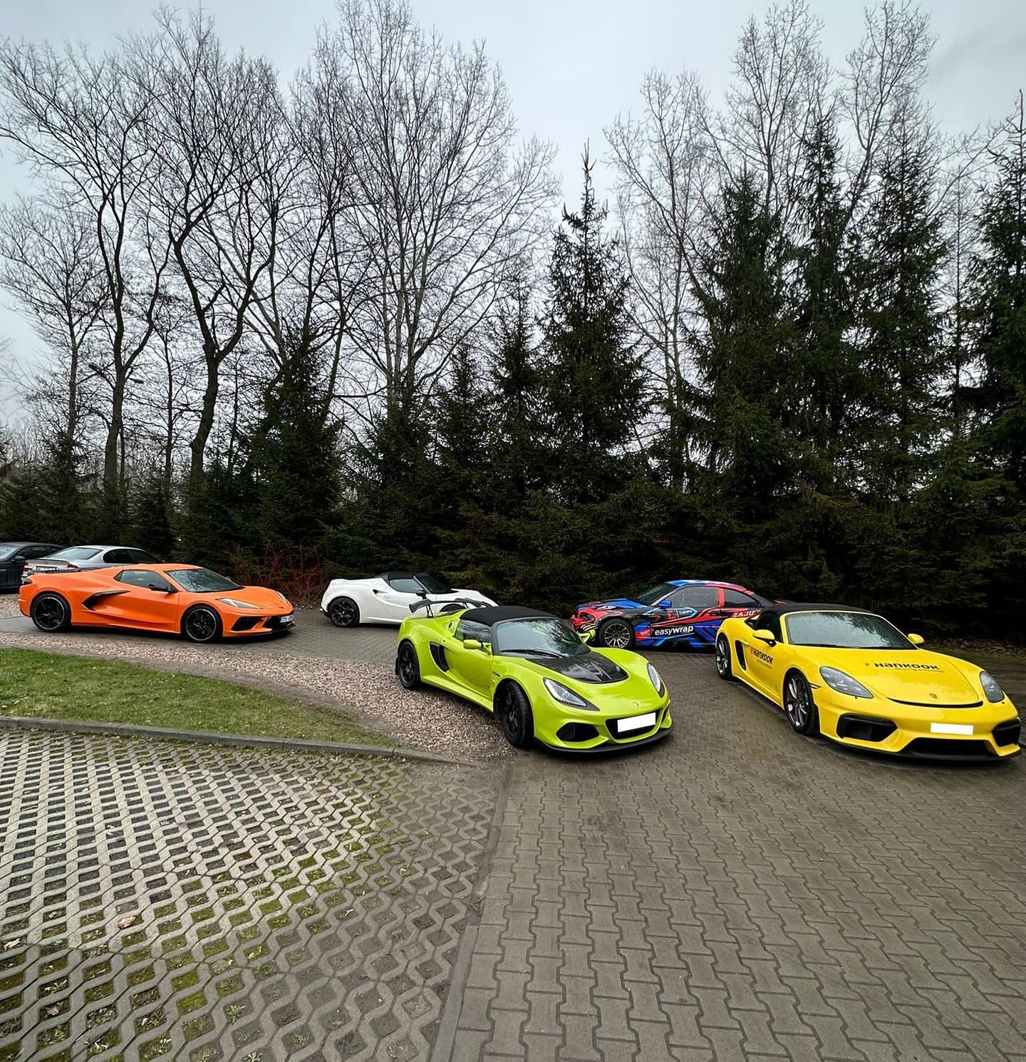 Kolorowe, sportowe auta (żółty Porsche, zielony Lotus) ustawione na parkingu z kostki, w tle drzewa. Jeden z samochodów ma okleinę EasyWrap.
