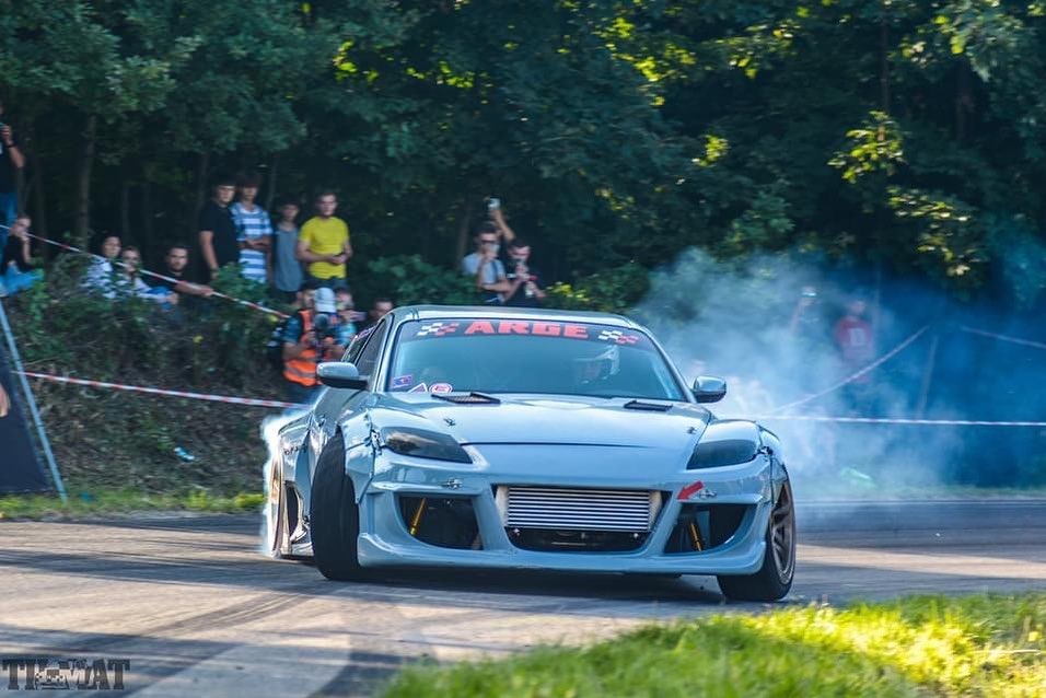 Błękitny, zmodyfikowany samochód sportowy w drifcie, z widoczną publicznością w tle i dymem z opon, na torze wyścigowym.