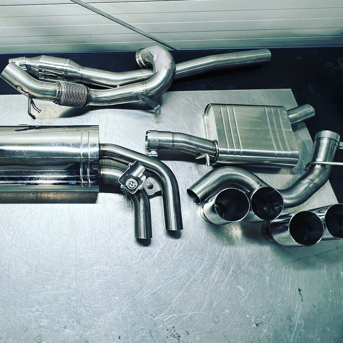 Kompletny, polerowany układ wydechowy ze stali nierdzewnej, z widocznymi tłumikami i rurami, leżący na metalowym stole warsztatowym. Układ gotowy do montażu.