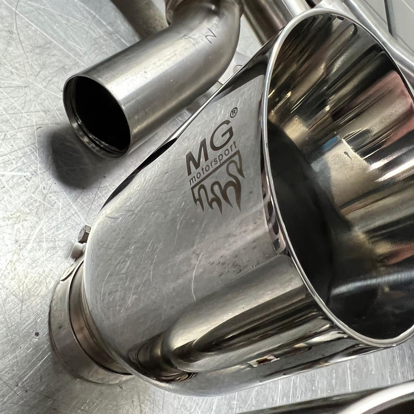 Polerowany, metalowy wydech samochodowy z logo MG Motorsport leży na metalowej powierzchni. Widoczne inne elementy wydechu w tle.