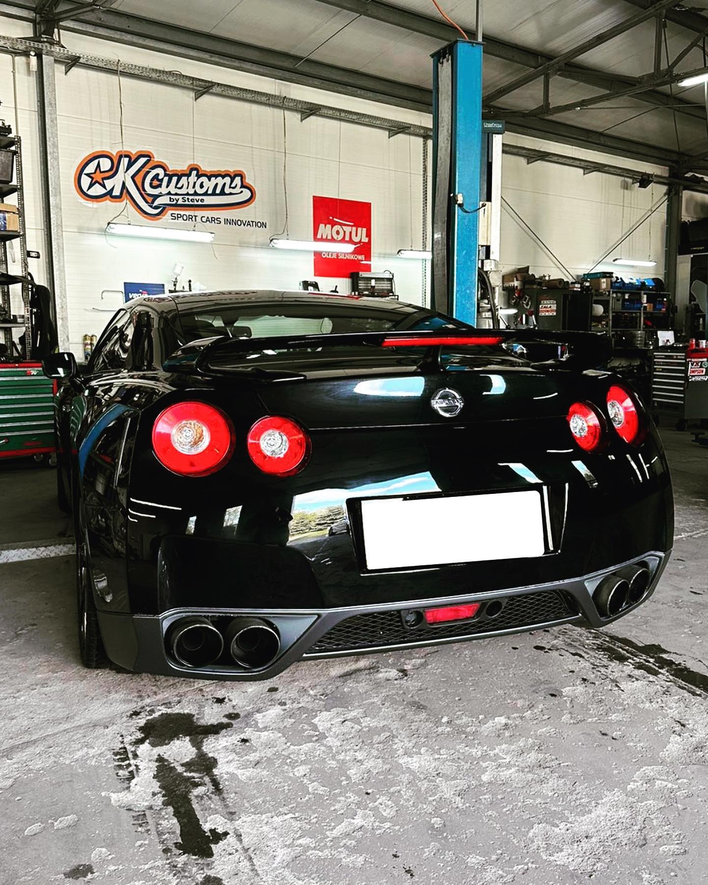 Czarny Nissan GT-R w warsztacie, widok z tyłu, z podwójnymi wydechami i czerwonymi światłami. W tle podnośnik i logo warsztatu.