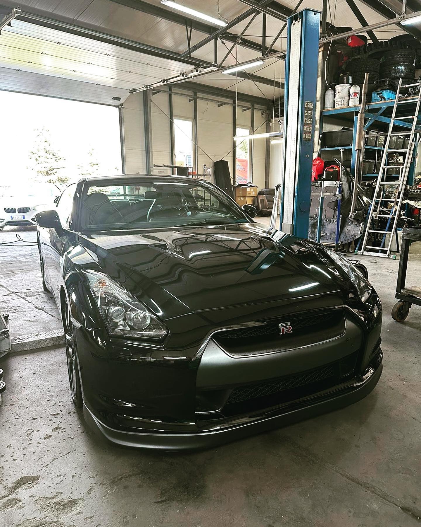 Czarny Nissan GT-R w warsztacie; widoczny podnośnik, regał z oponami i narzędziami. Ujęcie na przód auta, z detalami grilla i reflektorów.