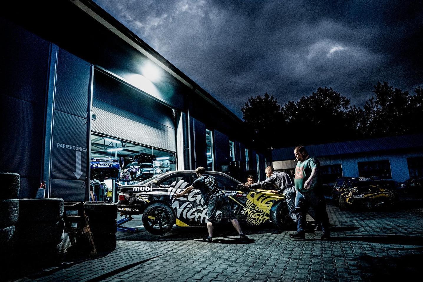 Czarny, oklejony samochód w warsztacie, z otwartą bramą, gdzie widać inne pojazdy. Grupa osób przy aucie, ciemne niebo w tle.