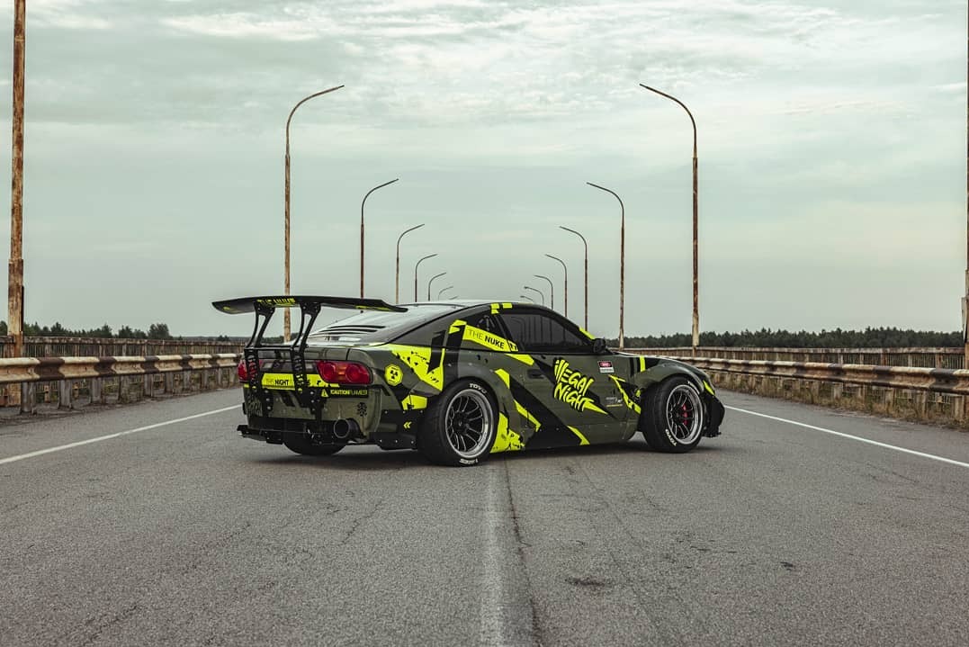 Tuningowany samochód z dużym spoilerem, w barwach khaki i neonowej zieleni, stoi na moście. Widok z tyłu, oświetlenie pochmurne.