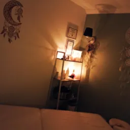 Przytulne wnętrze gabinetu masażu: łóżko do masażu, półka z certyfikatami i olejkami, dekoracje w stylu boho, ciepłe oświetlenie tworzące relaksującą atmosferę.