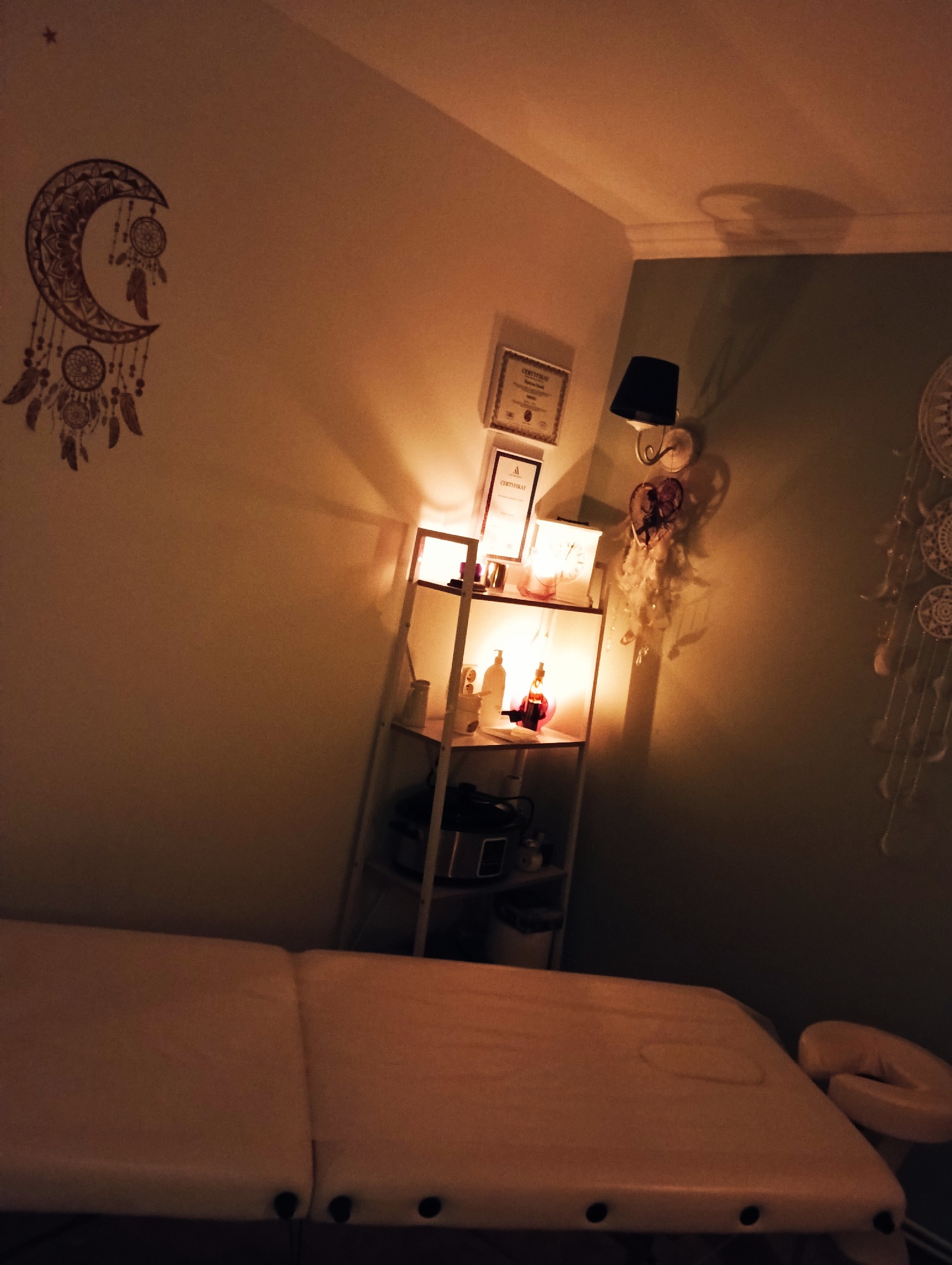 Przytulne wnętrze gabinetu masażu: łóżko do masażu, półka z certyfikatami i olejkami, dekoracje w stylu boho, ciepłe oświetlenie tworzące relaksującą atmosferę.