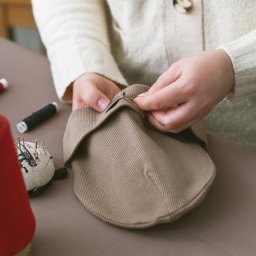 InUse sp z o o - Krawcowa ręcznie zszywa beżową czapkę na stole z akcesoriami do szycia: szpulki nici, poduszka z igłami. Jasne oświetlenie, ciepłe kolory.