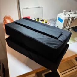 Dwa czarne, prostokątne pokrowce z tkaniny ułożone jeden na drugim na stole krawieckim, obok maszyna do szycia i lampa solna. Widoczne detale wykończenia.