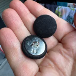 Dwa czarne, okrągłe guziki w dłoni, jeden z metalowym spodem z numerem 36. Tło rozmyte, widoczne ubrania. Detal krawiecki w zbliżeniu.