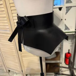 Czarny, skórzany pas biodrowy z kokardą, zaprezentowany na manekinie krawieckim. Widoczne detale szycia i wykończenia. Tło stanowią elementy wyposażenia pracowni.