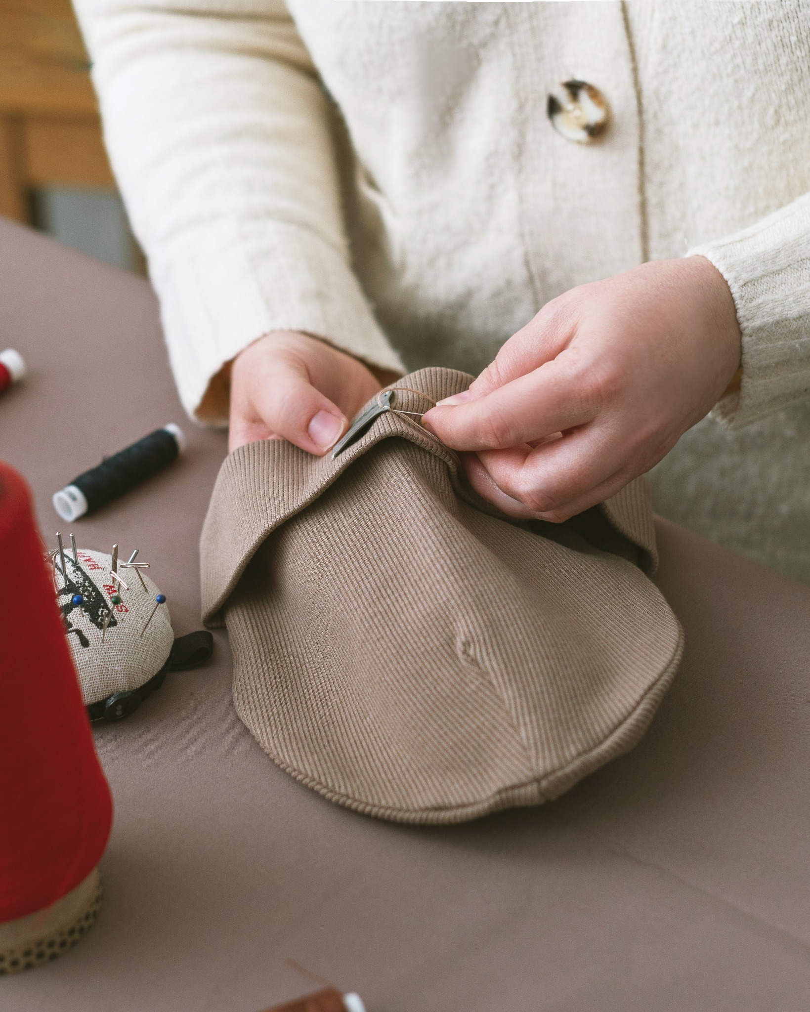 Krawcowa ręcznie zszywa beżową czapkę na stole z akcesoriami do szycia: szpulki nici, poduszka z igłami. Jasne oświetlenie, ciepłe kolory.