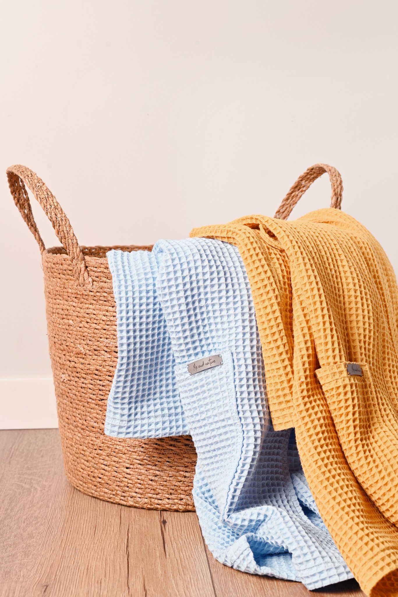 Koszyk z naturalnego materiału wypełniony błękitnym i żółtym, teksturowanym materiałem, prawdopodobnie ręcznikami lub szlafrokami z metkami.