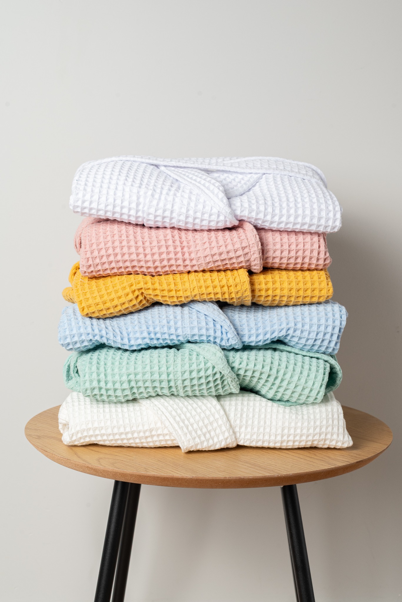 Stos ręcznie szytych szlafroków z waflowej bawełny w pastelowych kolorach (biały, różowy, żółty, niebieski, miętowy) ułożonych na okrągłym, drewnianym stoliku.