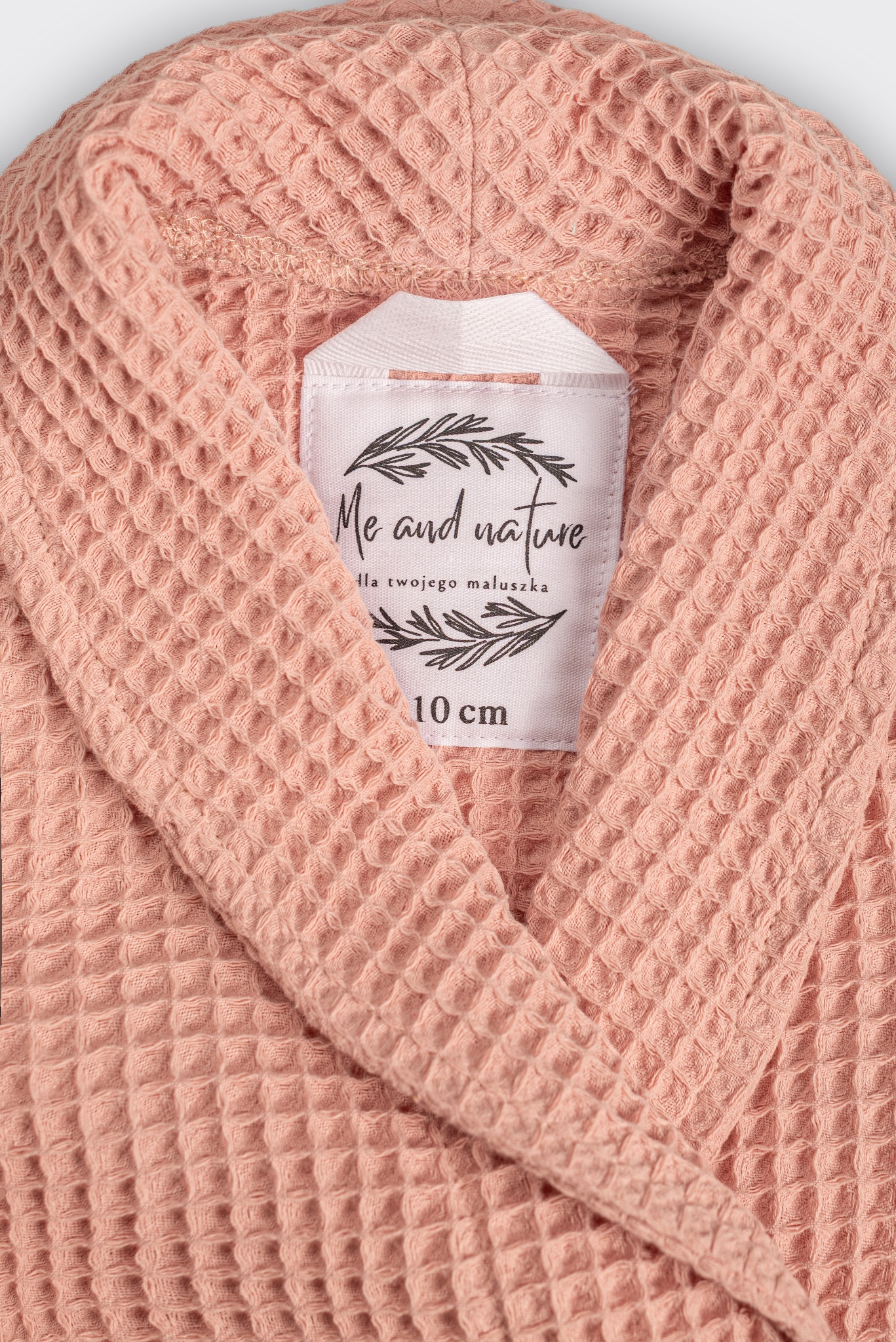 Różowy, wafelkowy szlafrok dziecięcy z białą metką 'Me and nature' i informacją o rozmiarze 10 cm. Ujęcie z bliska, prezentujące fakturę materiału i detale wykończenia.