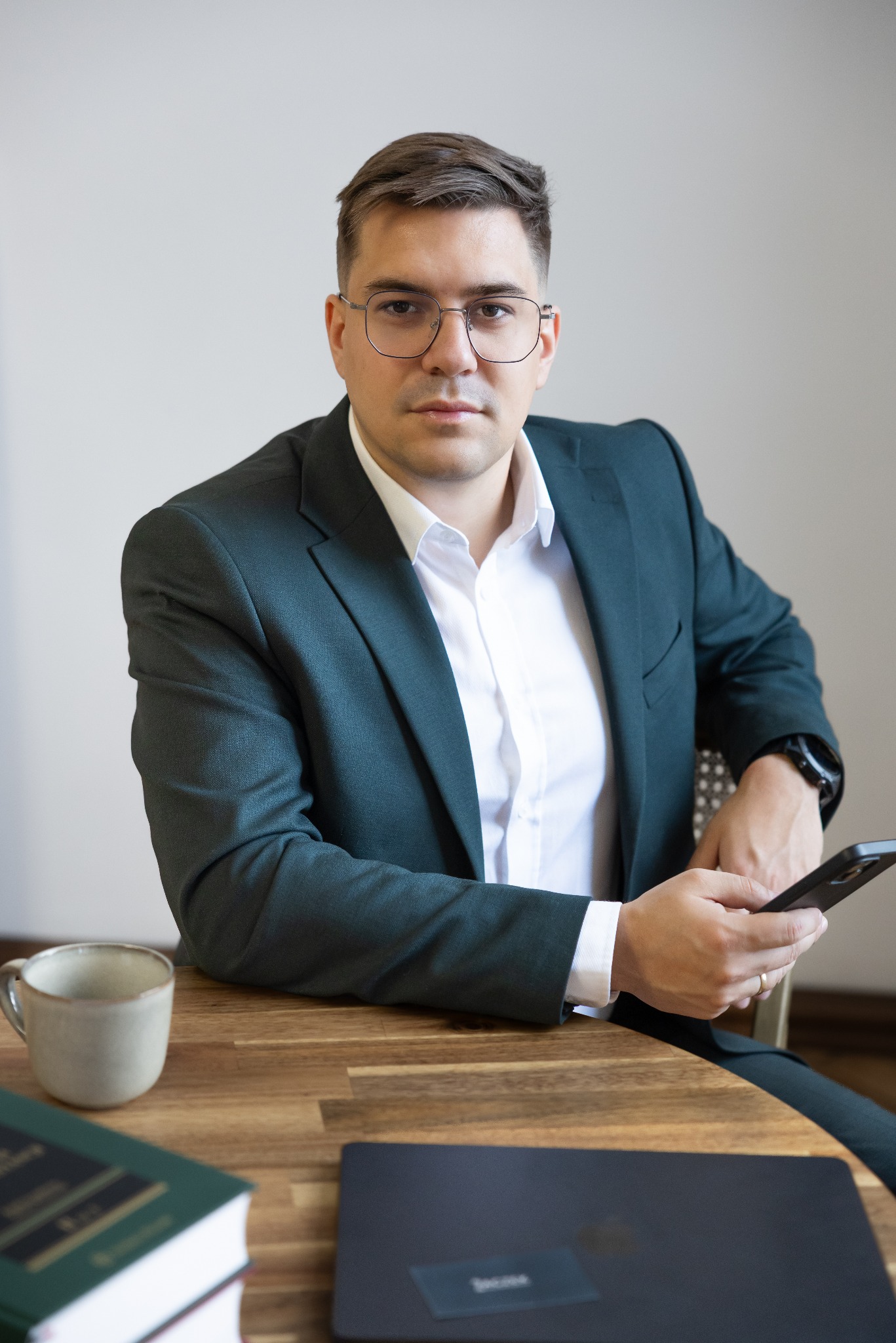 Mężczyzna w zielonym garniturze i okularach siedzi przy biurku z książkami, kubkiem i laptopem, trzymając telefon. Spokojny wyraz twarzy, profesjonalny wygląd.