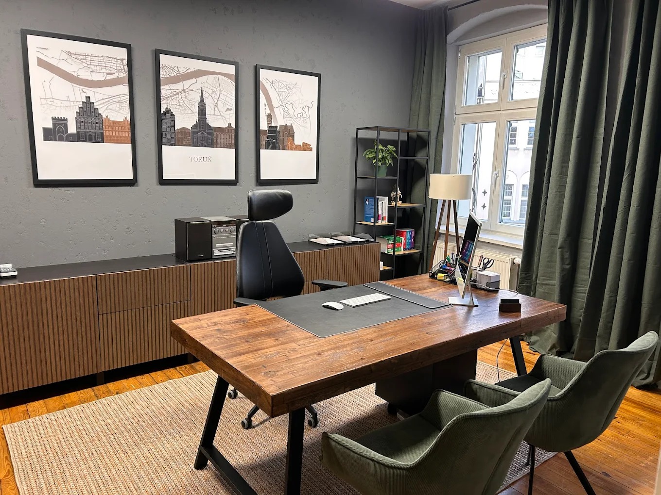 Nowoczesne biuro z drewnianym biurkiem, krzesłami i grafikami Torunia na ścianie. Stonowana kolorystyka, minimalistyczny design, widok na okno z zasłonami.