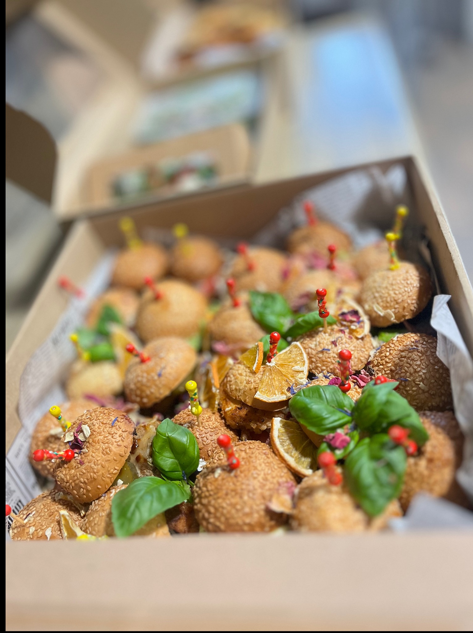 Zbliżenie na pudełko z mini burgerami na białym papierze, ozdobione bazylią i suszonymi pomarańczami, spięte dekoracyjnymi wykałaczkami. Catering.
