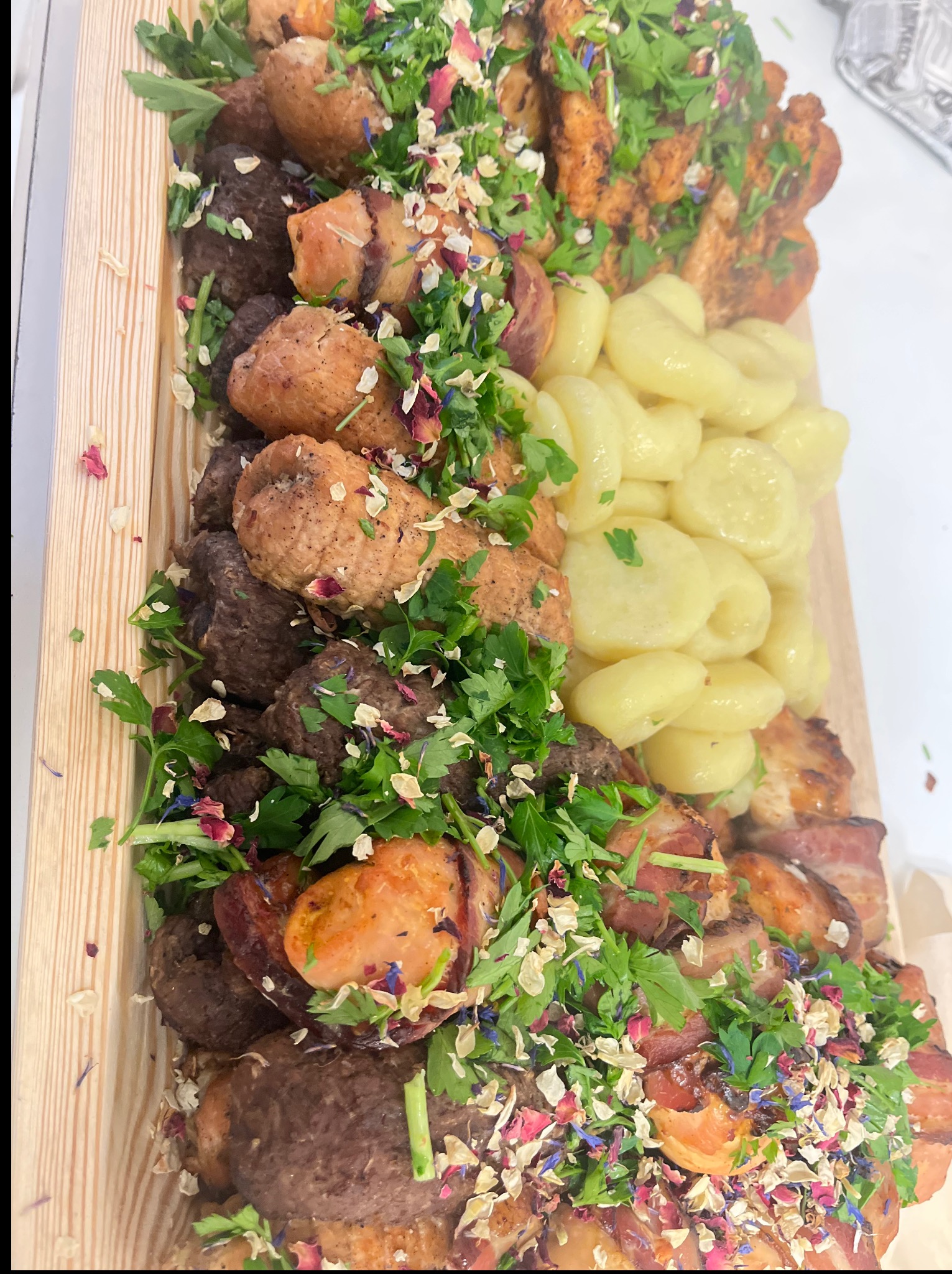 Dekoracyjna drewniana taca z różnorodnymi przekąskami: mięsami, ziemniakami i ziołami, idealna na catering firmowy lub świąteczny.