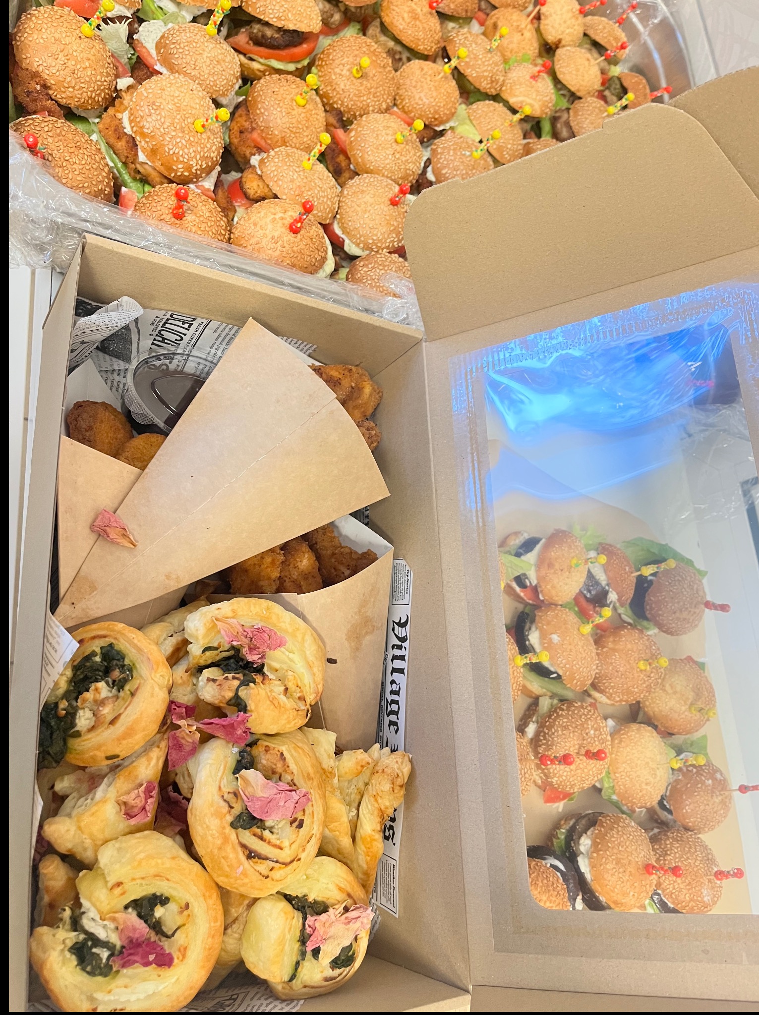 Zestaw cateringowy: mini burgery na srebrnej tacy i pudełka z przekąskami, w tym roladki ze szpinakiem i smażone kąski, udekorowane suszonymi płatkami róż.