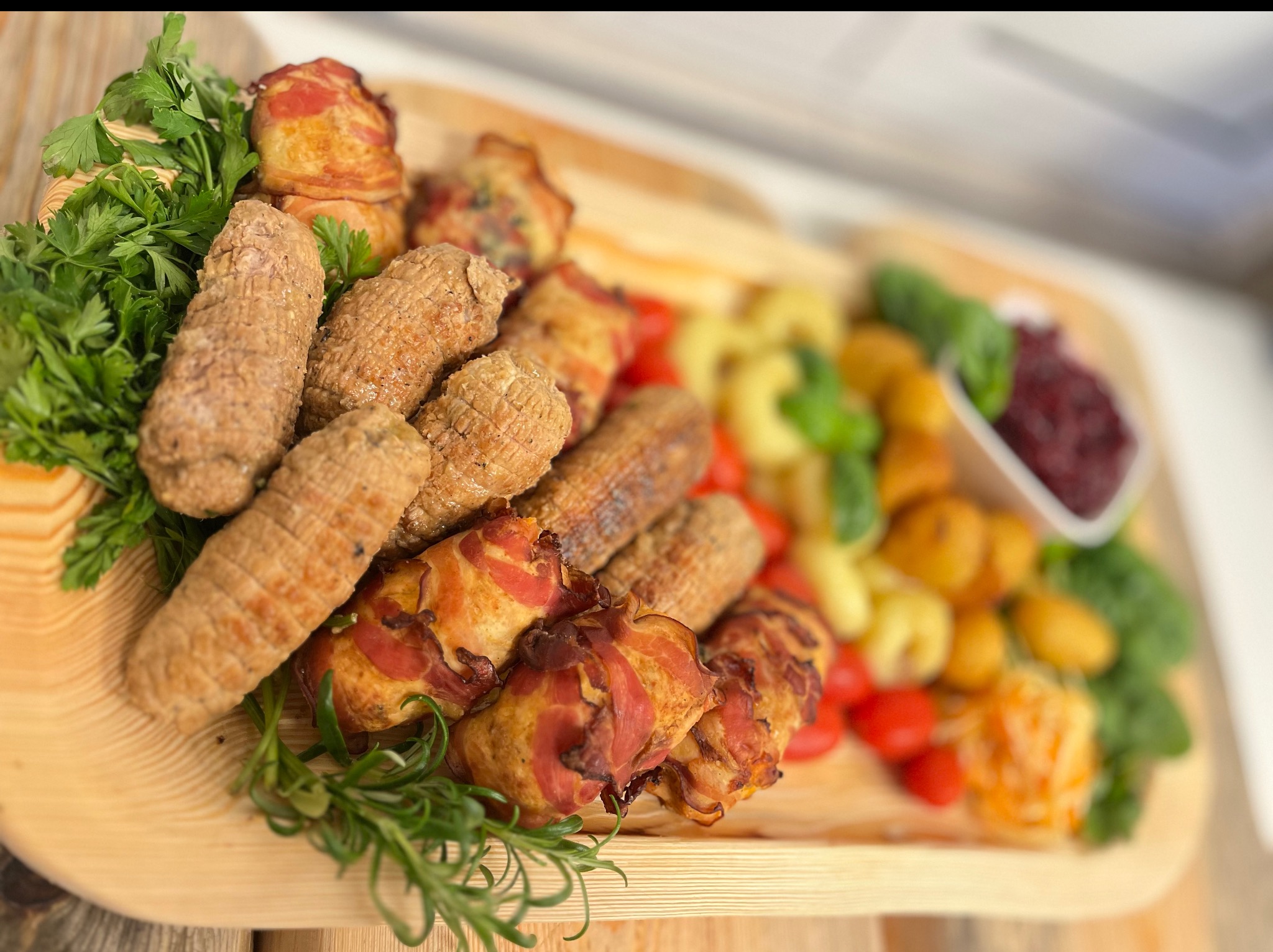 Drewniana taca z roladkami mięsnymi, zawijańcami z boczku, makaronem i sosem żurawinowym, udekorowana świeżymi ziołami. Elegancki catering na imprezę.