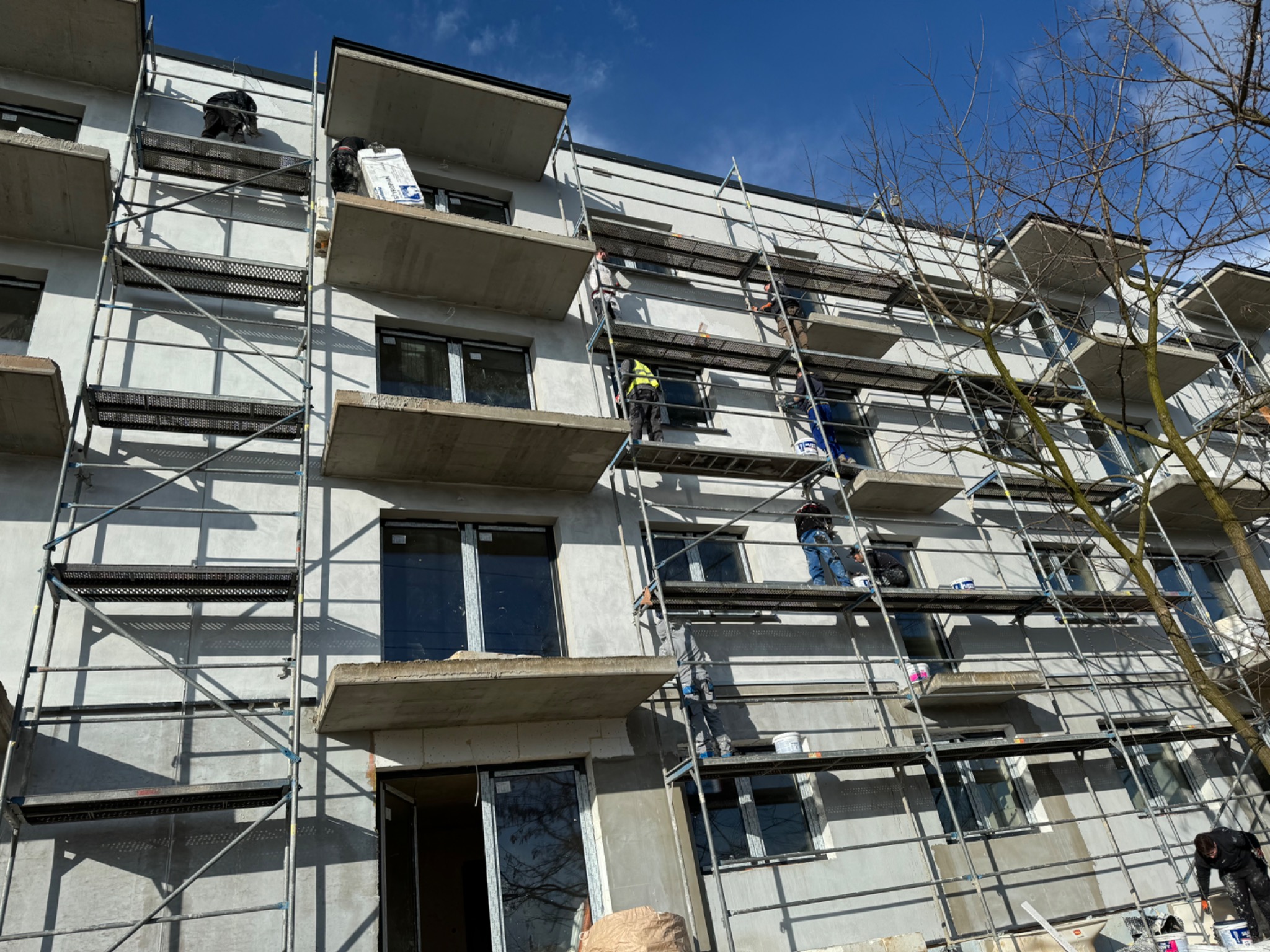 Prace na elewacji budynku z rusztowaniem. Widoczni pracownicy w trakcie wykonywania prac elewacyjnych. Świeży tynk i okna z folią zabezpieczającą.