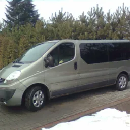 Szary bus Renault Traffic zaparkowany na brukowanym podjeździe, z ciemnymi szybami i widocznymi drzewami iglastymi w tle, w zimowej scenerii z resztkami śniegu.