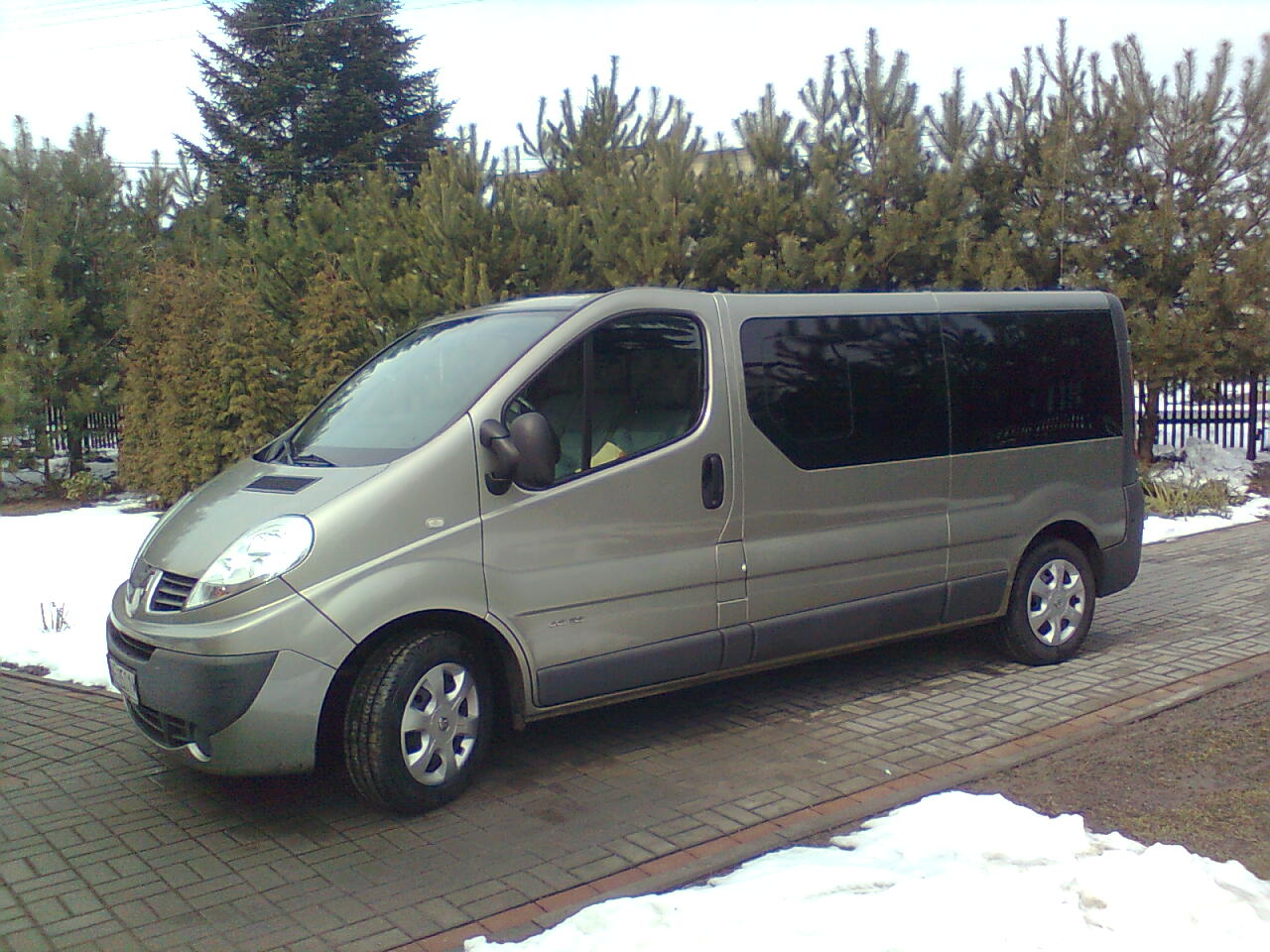 Szary bus Renault Traffic zaparkowany na brukowanym podjeździe, z ciemnymi szybami i widocznymi drzewami iglastymi w tle, w zimowej scenerii z resztkami śniegu.