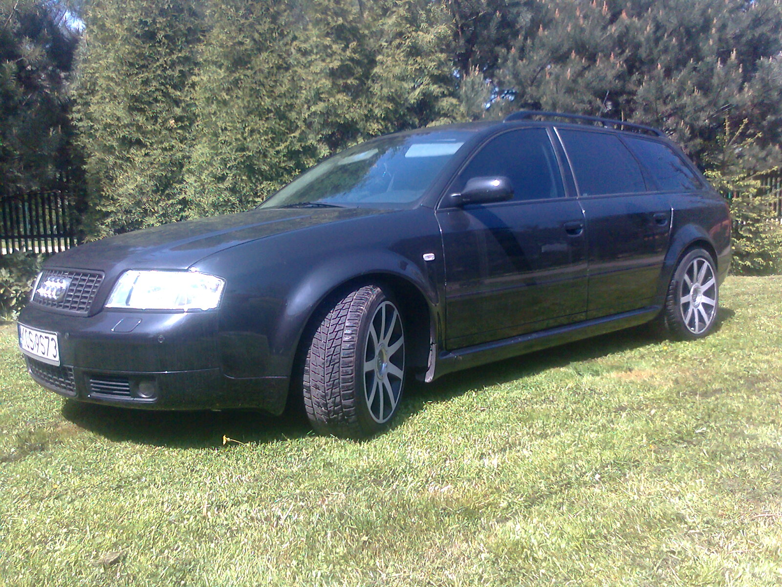 Ciemny samochód kombi marki Audi, zaparkowany na trawniku przed żywopłotem, widoczny z boku i przodu, z numerem rejestracyjnym na przednim zderzaku.