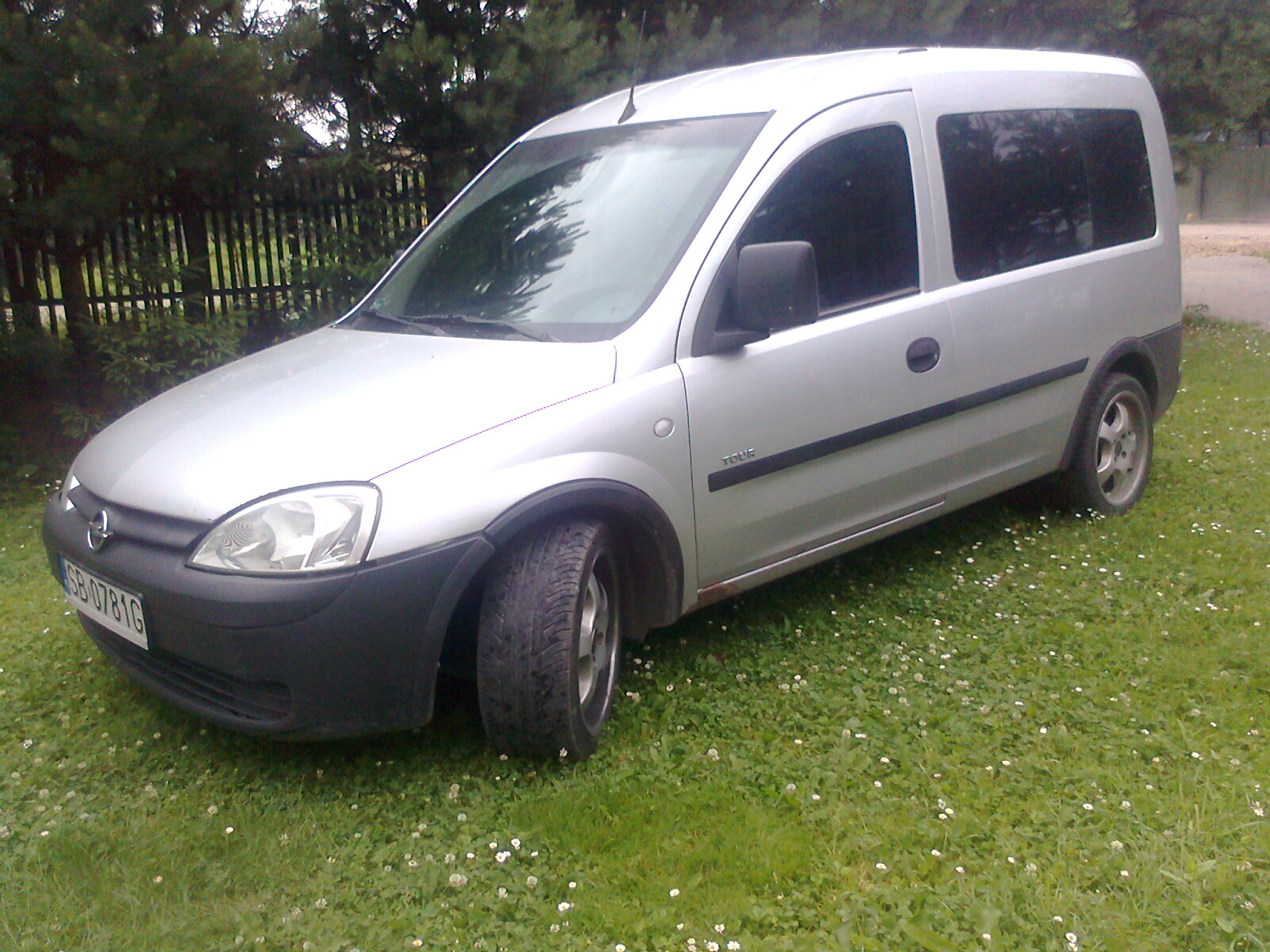 Srebrny Opel Combo Tour zaparkowany na trawniku z koniczyną, widoczny z boku, zarysowany próg i tablica rejestracyjna.
