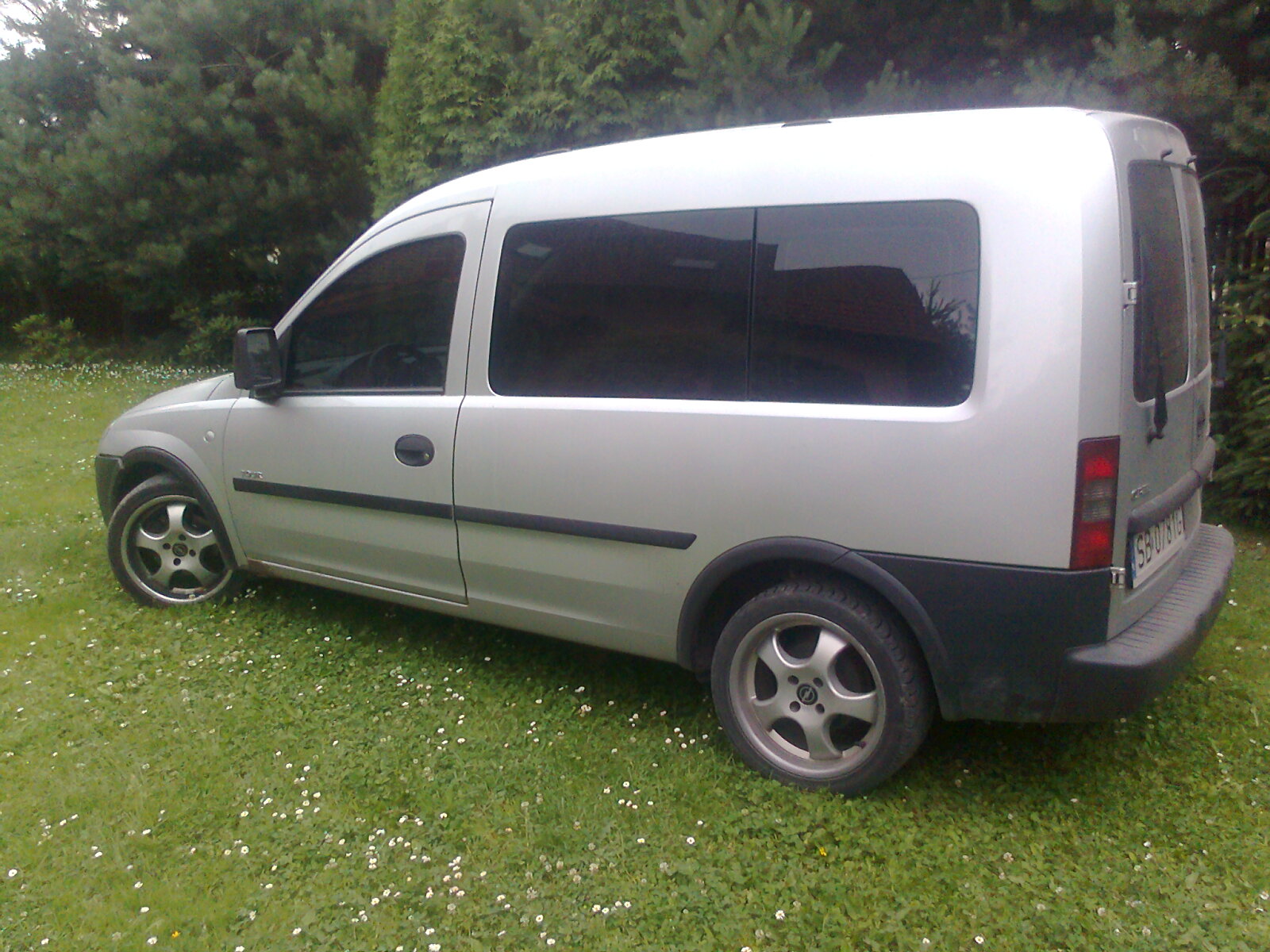 Srebrny samochód dostawczy Opel Combo z ciemnymi szybami, zaparkowany na trawniku z koniczyną, widok z boku.
