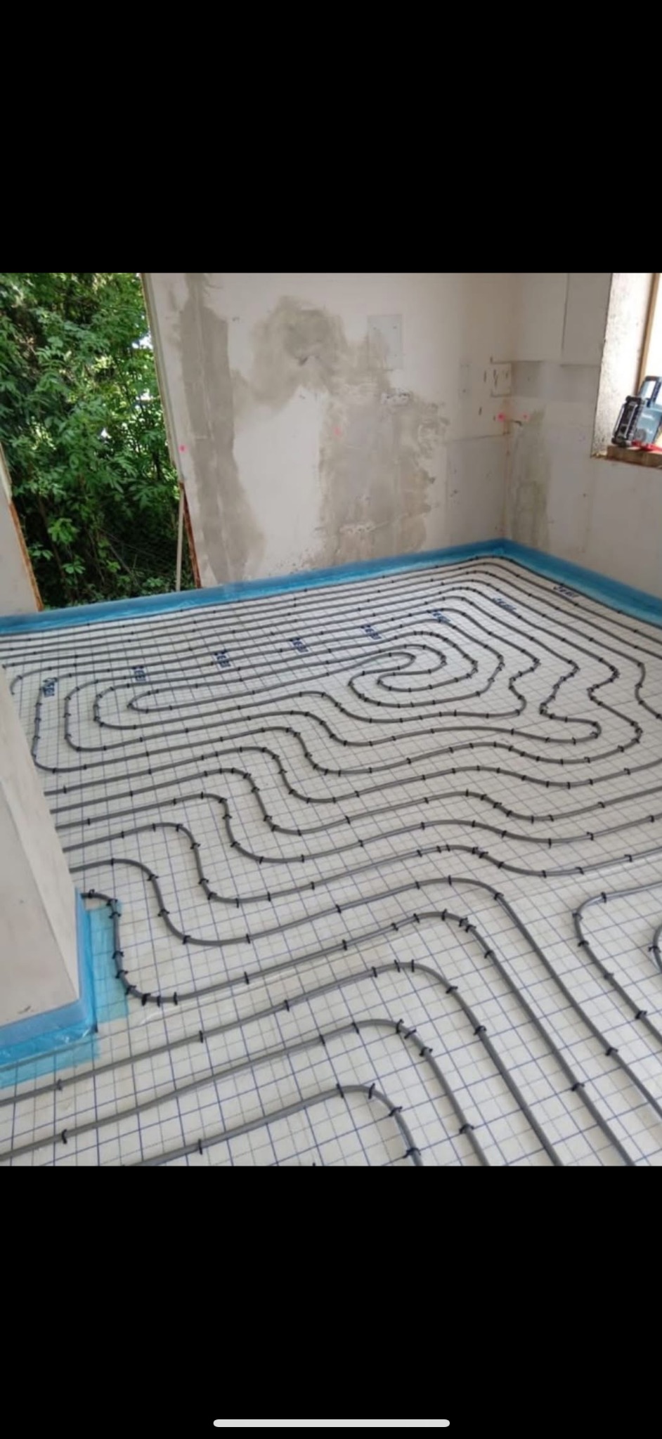 Szare rury ogrzewania podłogowego ułożone na białej macie w surowym wnętrzu z oknem i widokiem na zieleń. Niebieska folia zabezpiecza ściany.