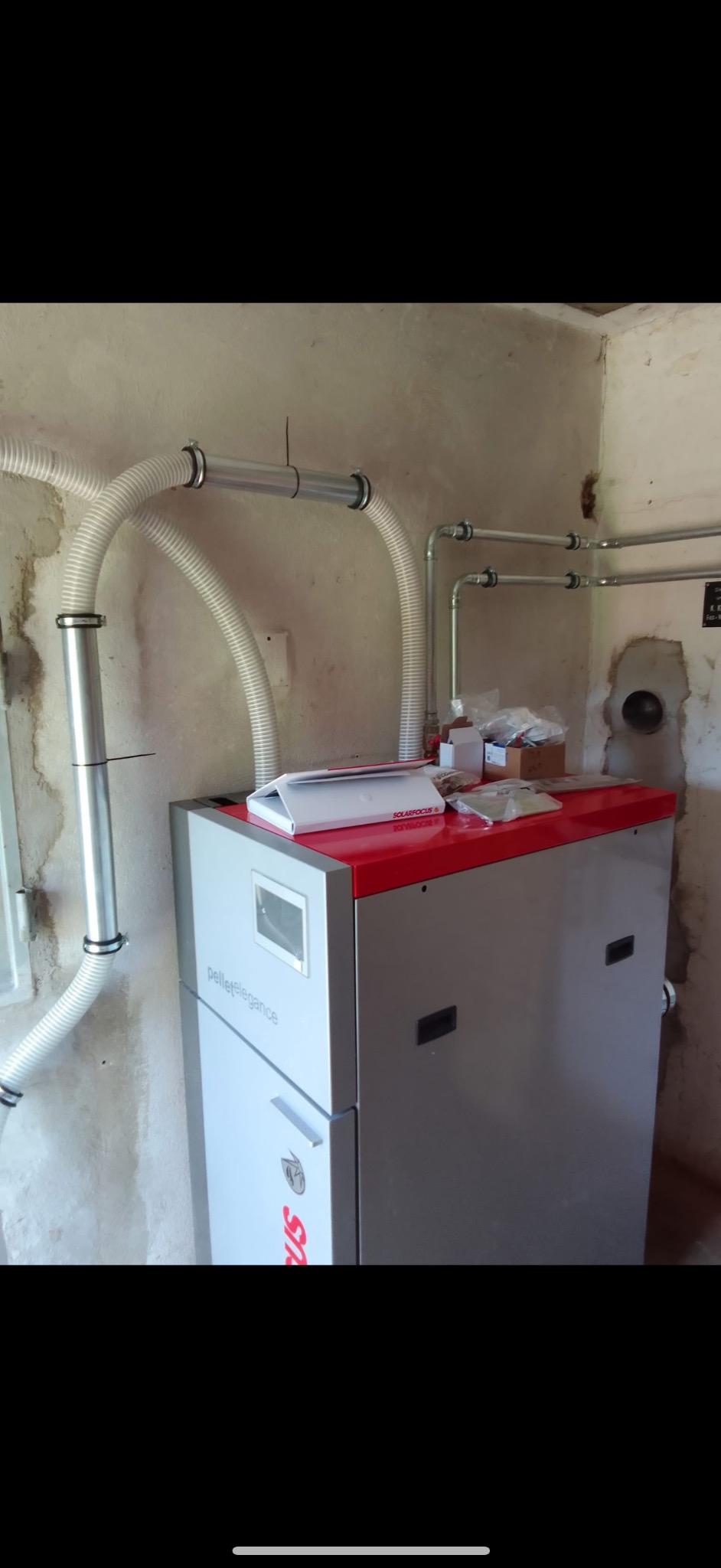 Szary piec pellet elegance z czerwonym blatem, podłączony do instalacji z rur aluminiowych i białych, karbowanych przewodów w surowym wnętrzu.