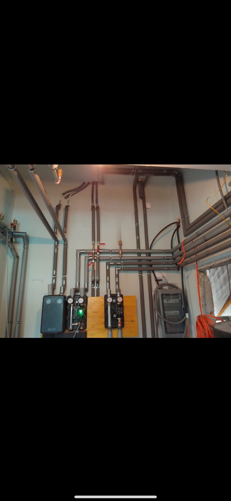 Szare rury instalacji grzewczej z zaworami i manometrami na ścianie. Widoczne pompy obiegowe, skrzynka elektryczna i orurowanie pod sufitem.