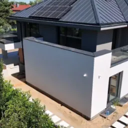 Nowoczesny dom z panelami słonecznymi na dachu, dużymi oknami i szklanymi balustradami balkonów. Widok z góry, otoczenie w trakcie prac wykończeniowych.