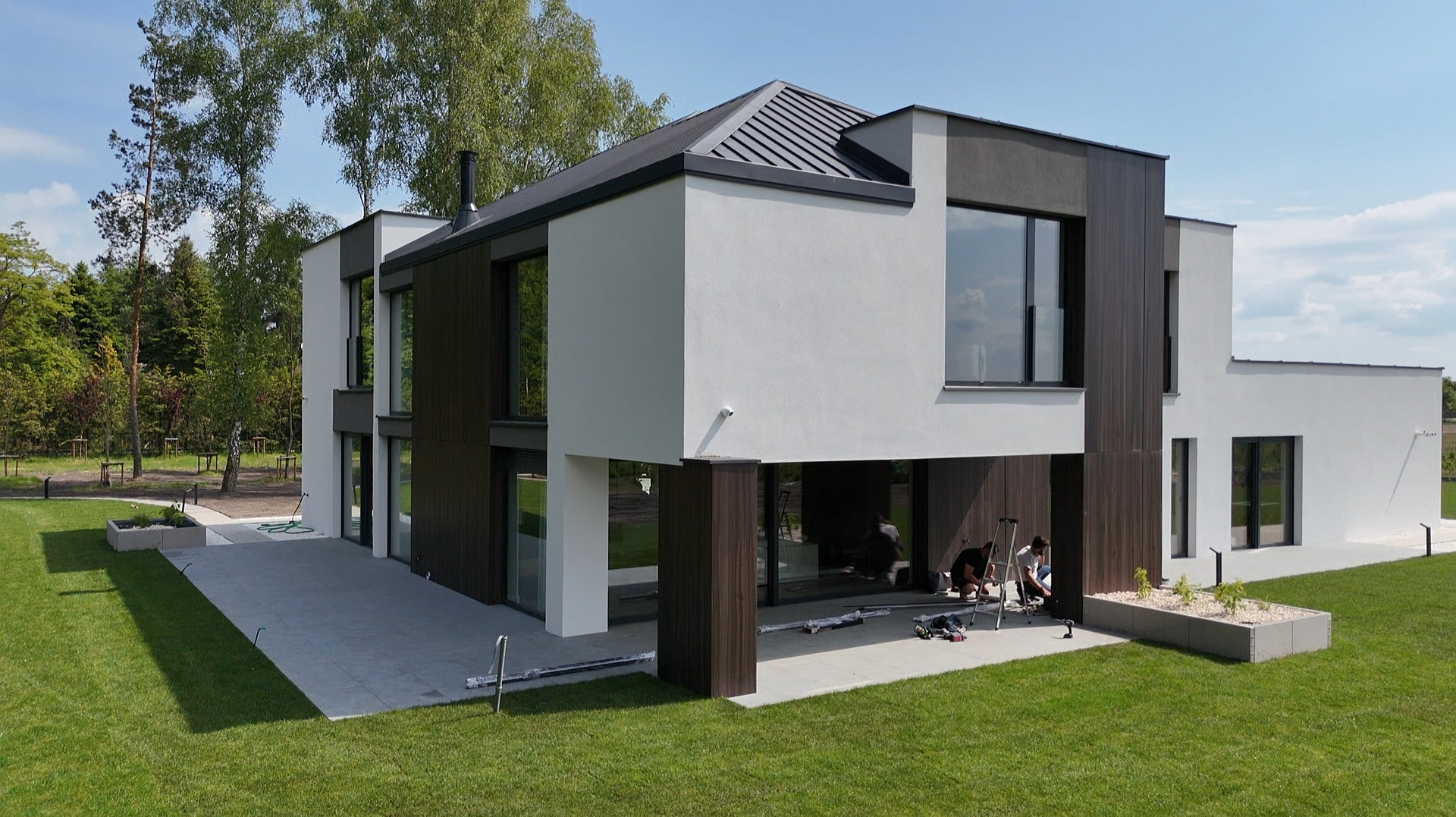 Nowoczesny dom z dużymi przeszkleniami i ciemnymi ramami okiennymi, widok z zewnątrz na taras z pracującymi osobami. Minimalistyczna architektura i zadbany trawnik.