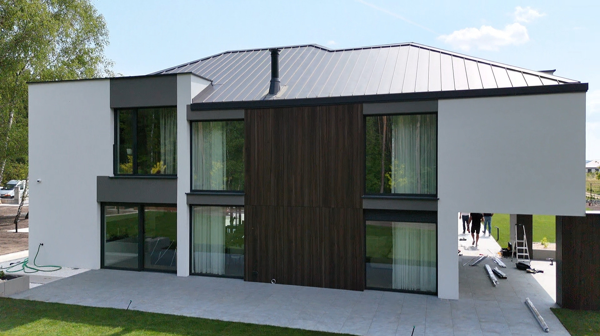 Nowoczesny dom z aluminiowymi oknami w grafitowych ramach, drewnianą fasadą i metalowym dachem. Na tarasie narzędzia i ekipa montażowa.