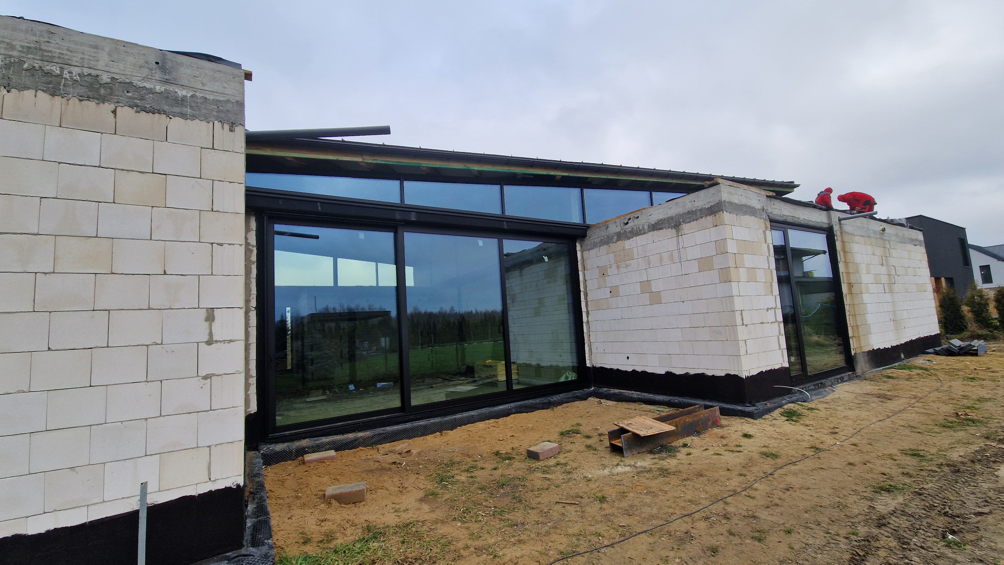 Montaż panoramicznych przeszkleń w surowym budynku. Widoczne detale konstrukcyjne i pracownicy na dachu. Duże okna z czarnymi ramami odbijają krajobraz.
