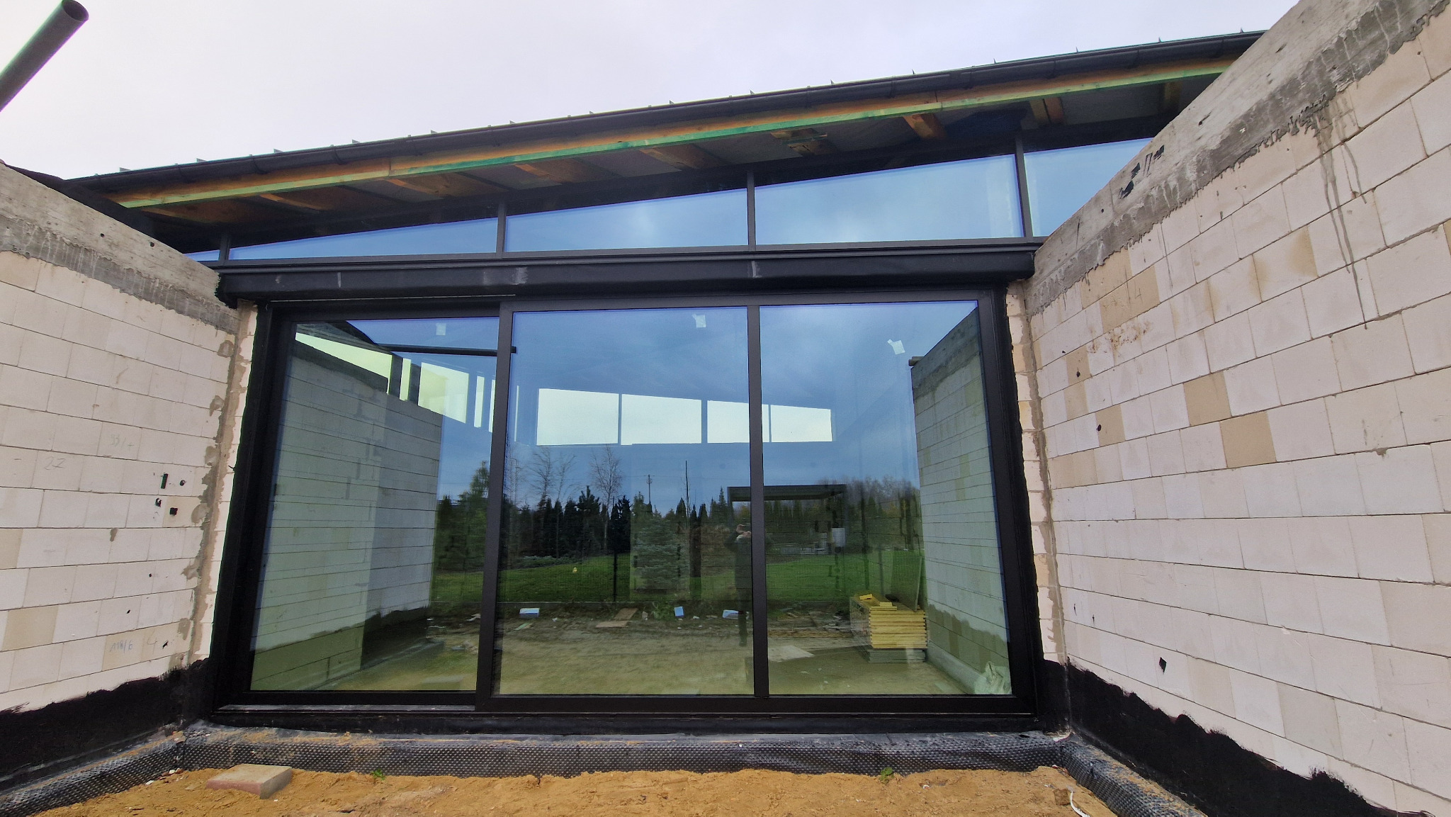 Duże, przesuwne okno z czarną ramą w surowym stanie budynku. Widoczna izolacja przeciwwilgociowa i pustaki. W tle zieleń ogrodu odbija się w szybie.